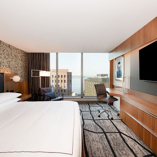 A modern hotel room features a king-sized bed, large TV, desk with chair, contemporary decor, and a window view of city buildings.
