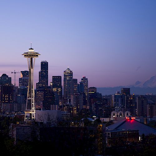 The image shows a city skyline at dusk, featuring a prominent tower and Mount Rainier in the background amidst a clear evening sky.