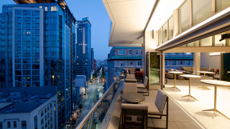 A modern rooftop terrace at dusk, furnished with tables and chairs, overlooking a cityscape with tall buildings and illuminated streets.