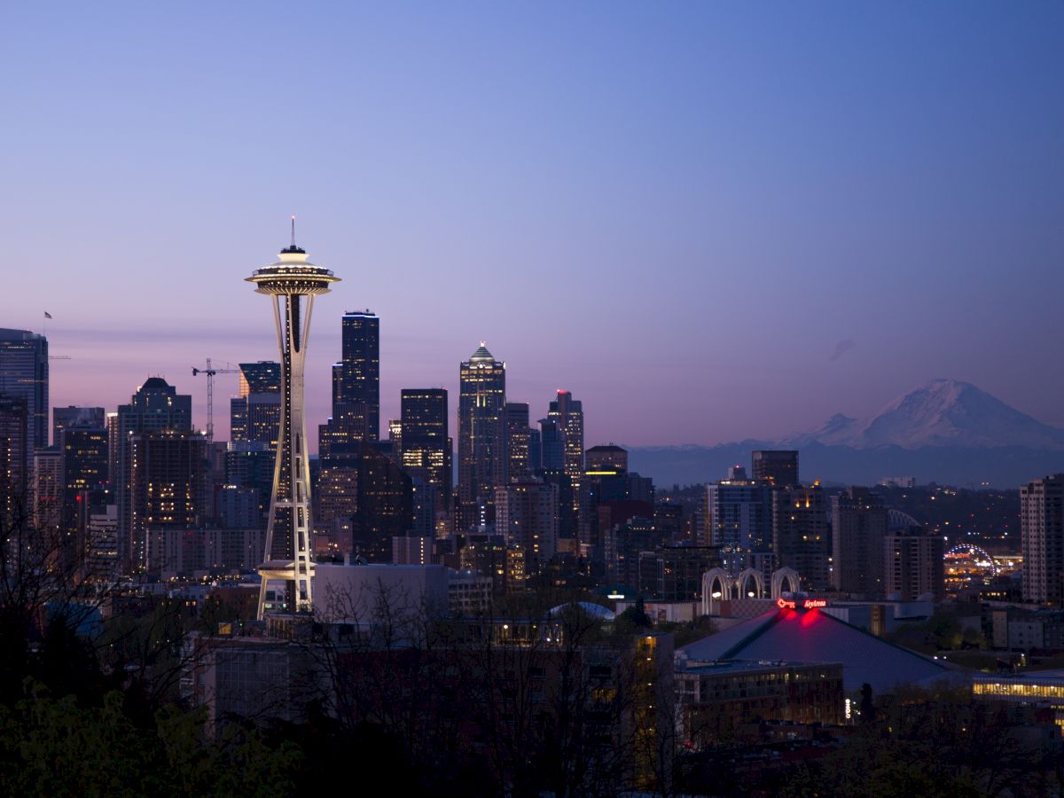 A twilight cityscape featuring a skyline with the iconic Space Needle, surrounded by various high-rise buildings and a distant mountain in the background.
