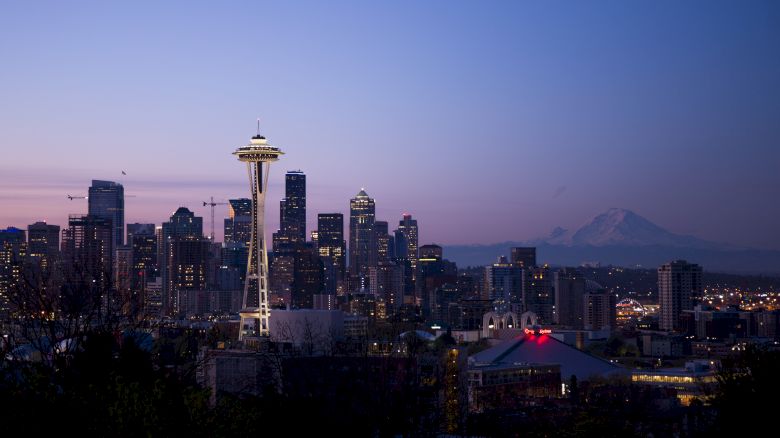 A twilight cityscape featuring a skyline with the iconic Space Needle, surrounded by various high-rise buildings and a distant mountain in the background.