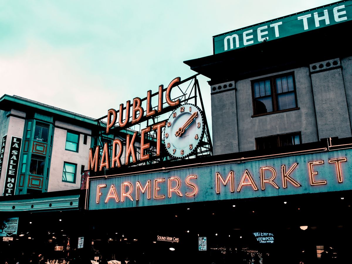 The image shows a well-known public market with neon signs reading "Public Market" and "Farmers Market," featuring a clock and urban buildings.
