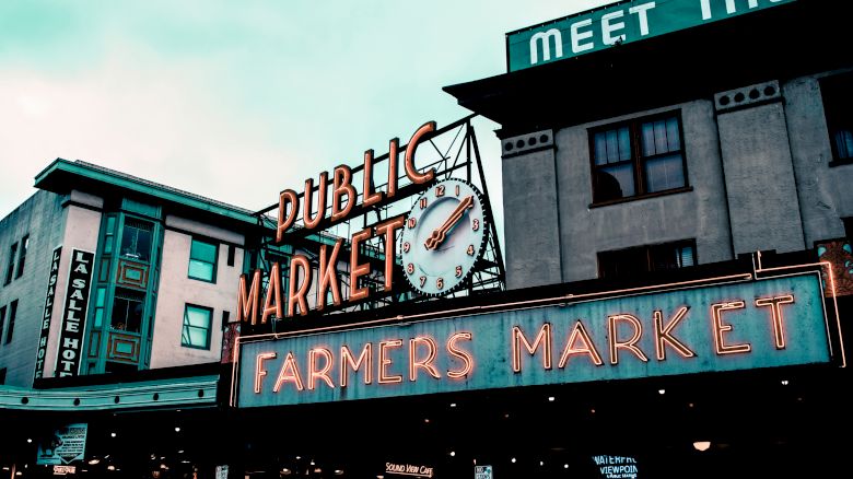 The image shows a well-known public market with neon signs reading "Public Market" and "Farmers Market," featuring a clock and urban buildings.