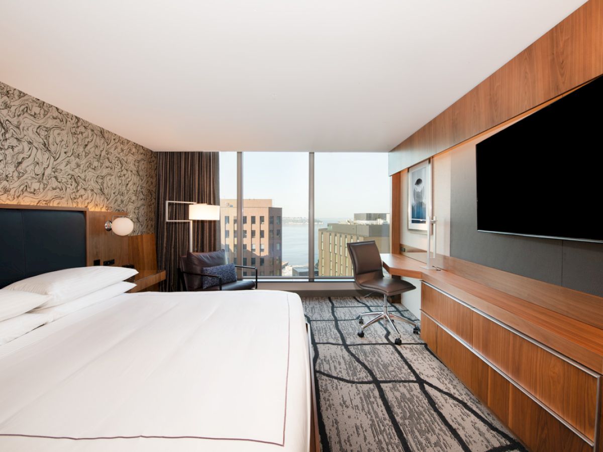 A modern hotel room with a large bed, desk with chair, flat-screen TV, and floor-to-ceiling windows offering a view of nearby buildings and water.