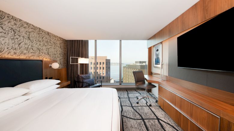 A modern hotel room with a large bed, desk with chair, flat-screen TV, and floor-to-ceiling windows offering a view of nearby buildings and water.