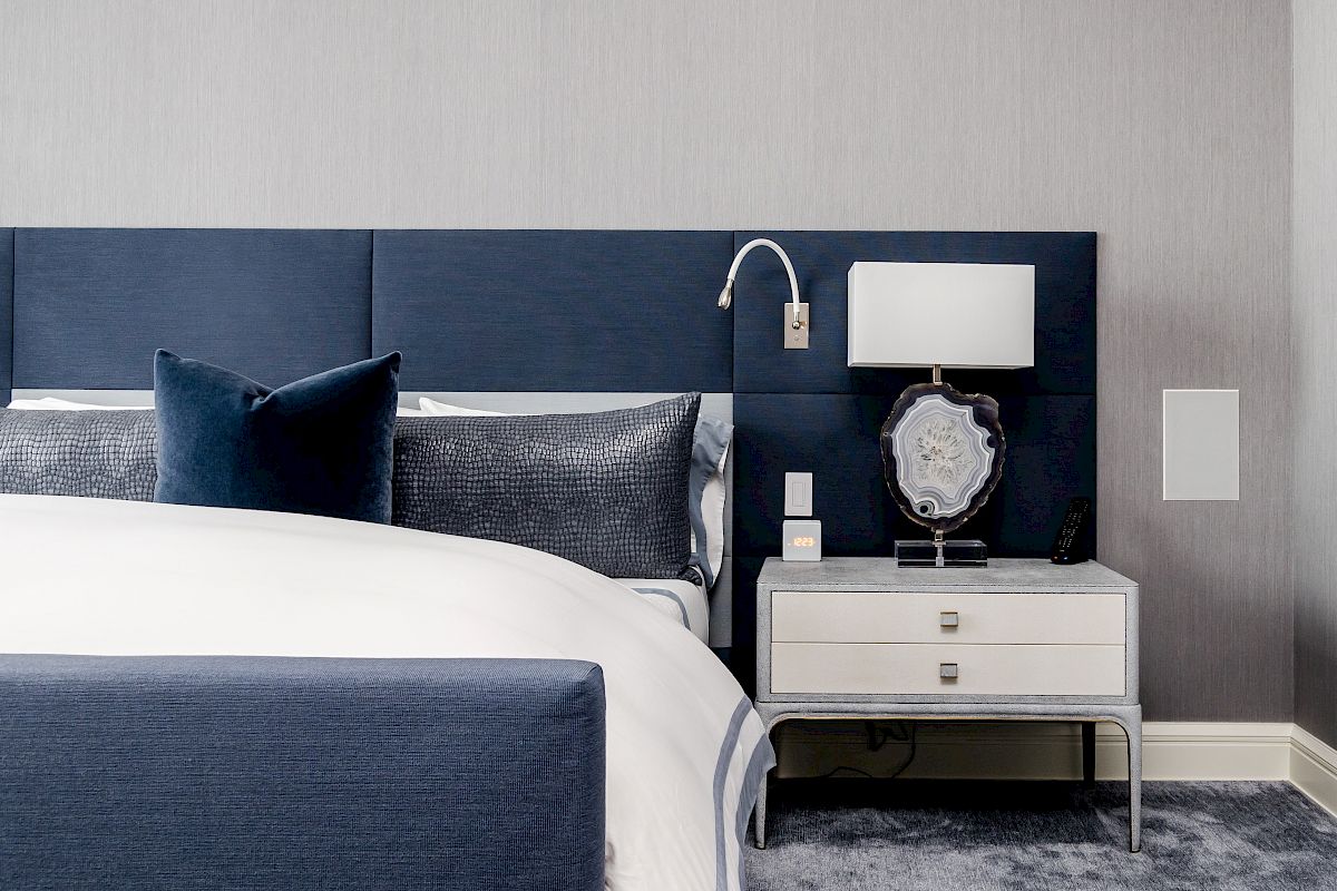 A modern bedroom features a navy blue headboard, white and blue bedding, a silver nightstand with two drawers, a lamp, and a decorative object.