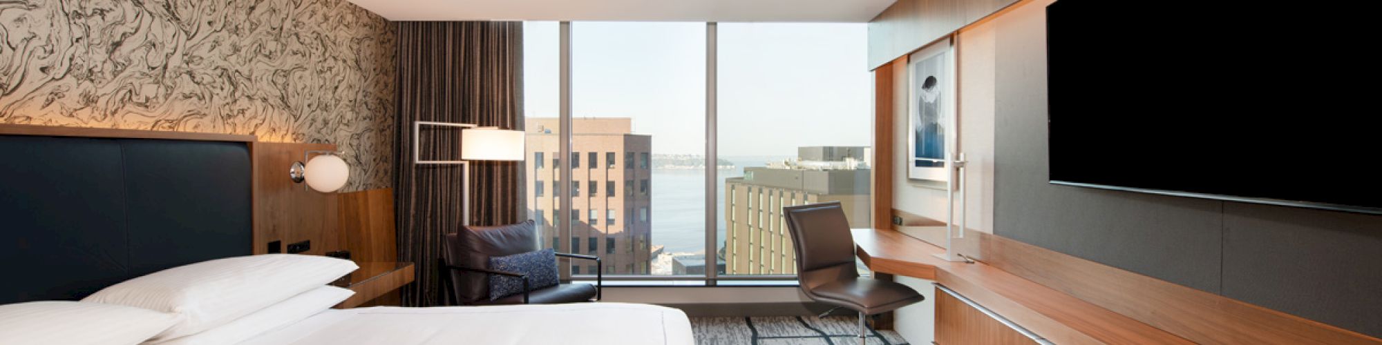 A modern hotel room features a bed, a large TV, a desk with a chair, and a window with a city view.